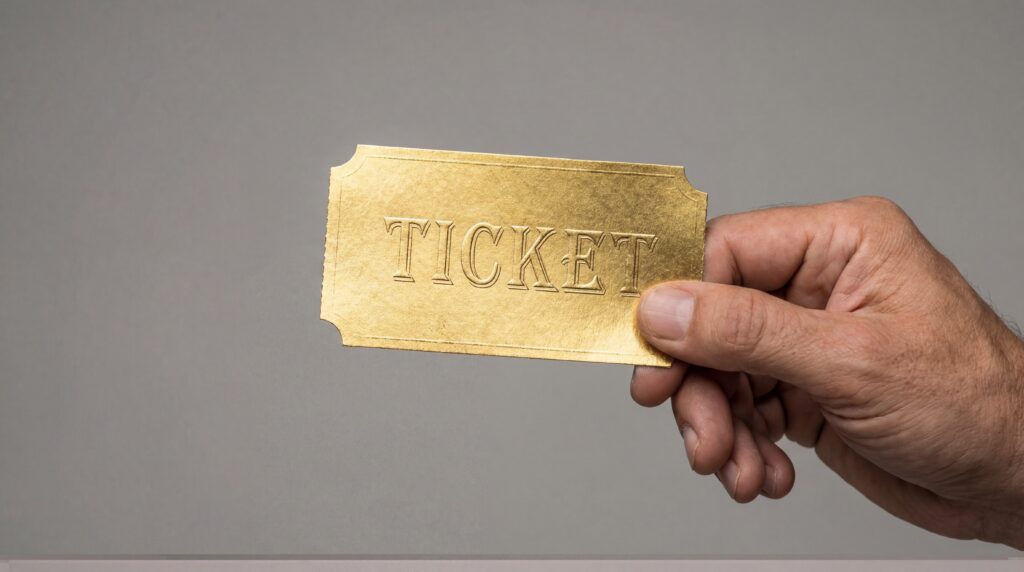 Hand holds a gold ticket with the word ticket embossed on its surface in a simple background setting